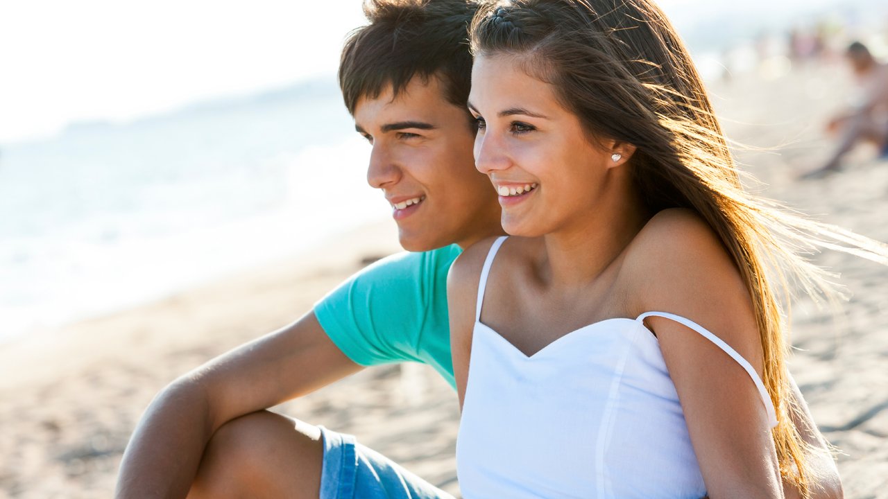 Cute teen couple sitting together on beach at sunset.