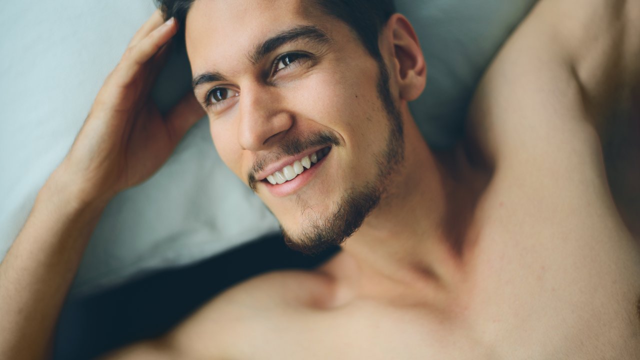 Handsome man lying on bed, happy smile waking up in the morning, attractive guy smiling in bedroom at home, sexy young male model
