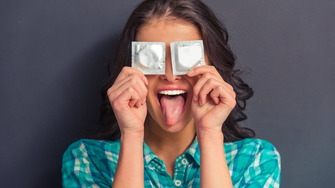 Portrait of attractive girl covering eyes with condoms and showing her tongue, against dark background