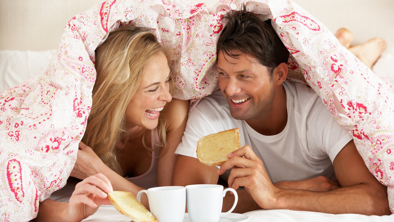 Couple Snuggled Under Duvet Eating Breakfast Smiling
