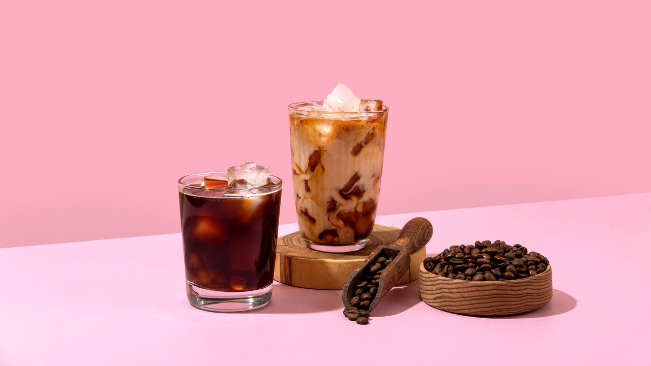 Ice coffee in a tall glass with cream poured over and coffee beans. Set with different types of coffee drinks on a pink table.