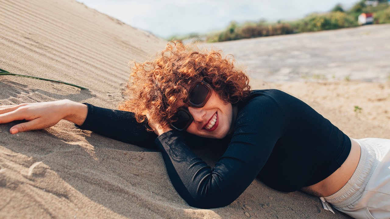 Close up shot of a beautiful young Caucasian woman with curly hair wearing a black long sleeve crop T-shirt and sunglasses while lying on her belly on a sand dune in a desert.