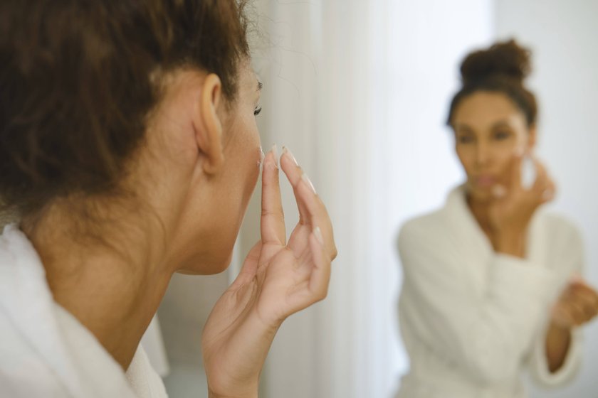 Eine Frau in einem weißen Bademantel cremt ihr Gesicht ein, während sie sich im Badezimmerspiegel betrachtet. Eine Frau in einem weißen Bademantel cremt ihr Gesicht ein, während sie sich im Badezimmerspiegel betrachtet.