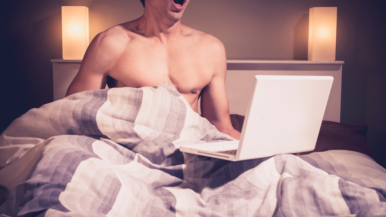 Young man is sitting in bed and watching pornography on his laptop