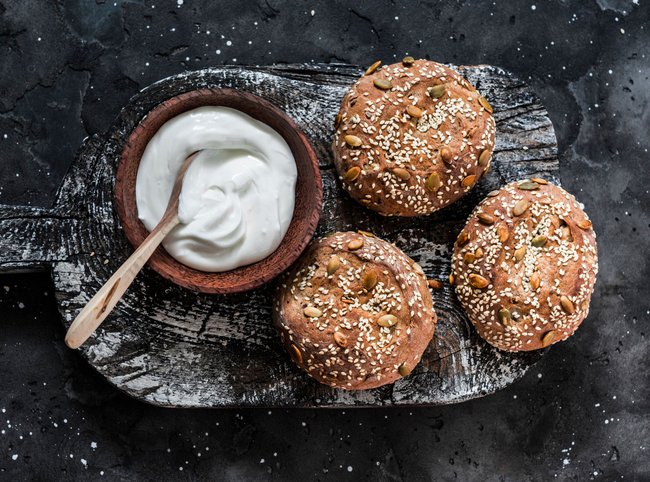 Multigrain multiseed fitness buns and greek yogurt on rustic wooden chopping board on dark background, top view Multigrain multiseed fitness buns and greek yogurt on rustic wooden chopping board on dark background, top view