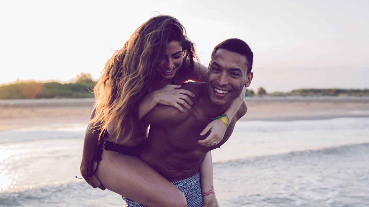 Young happy female tourist riding piggyback on African American boyfriend against wavy ocean during honeymoon Copyright: xVictorxGonzalezx