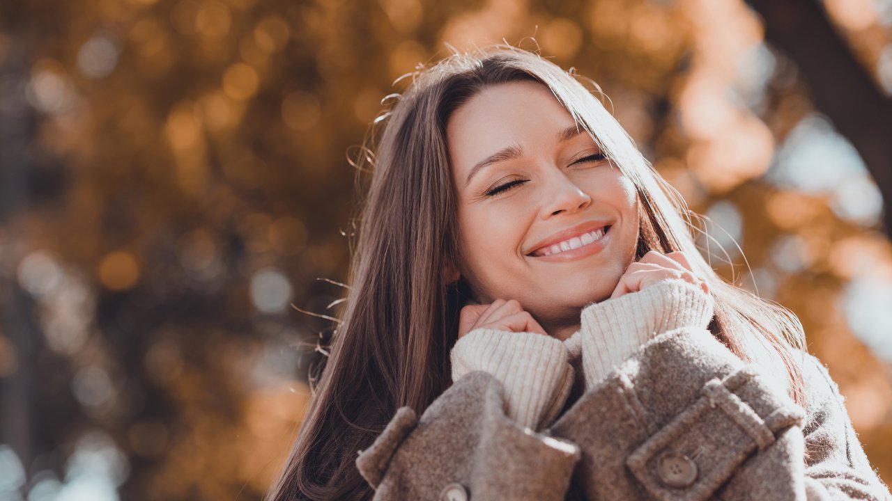 Eine lächelnde Frau ist im Herbst draußen und sie trägt einen braunen Mantel und einen hellen Schal. Eine lächelnde Frau ist im Herbst draußen und sie trägt einen braunen Mantel und einen hellen Schal.