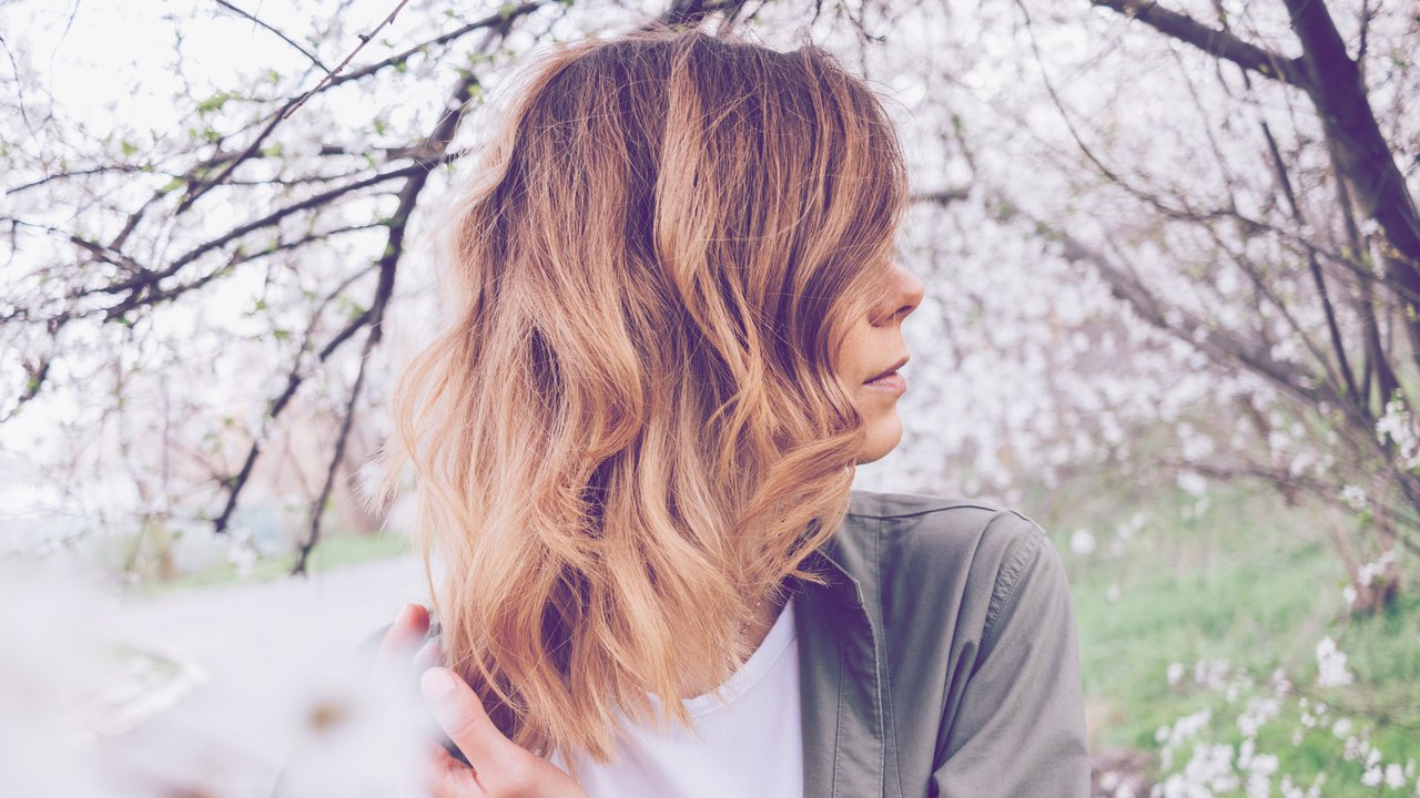 Side view close-up of female hairstyle long wavy bob. Young woman stands among blooming spring tree and touching her hair.