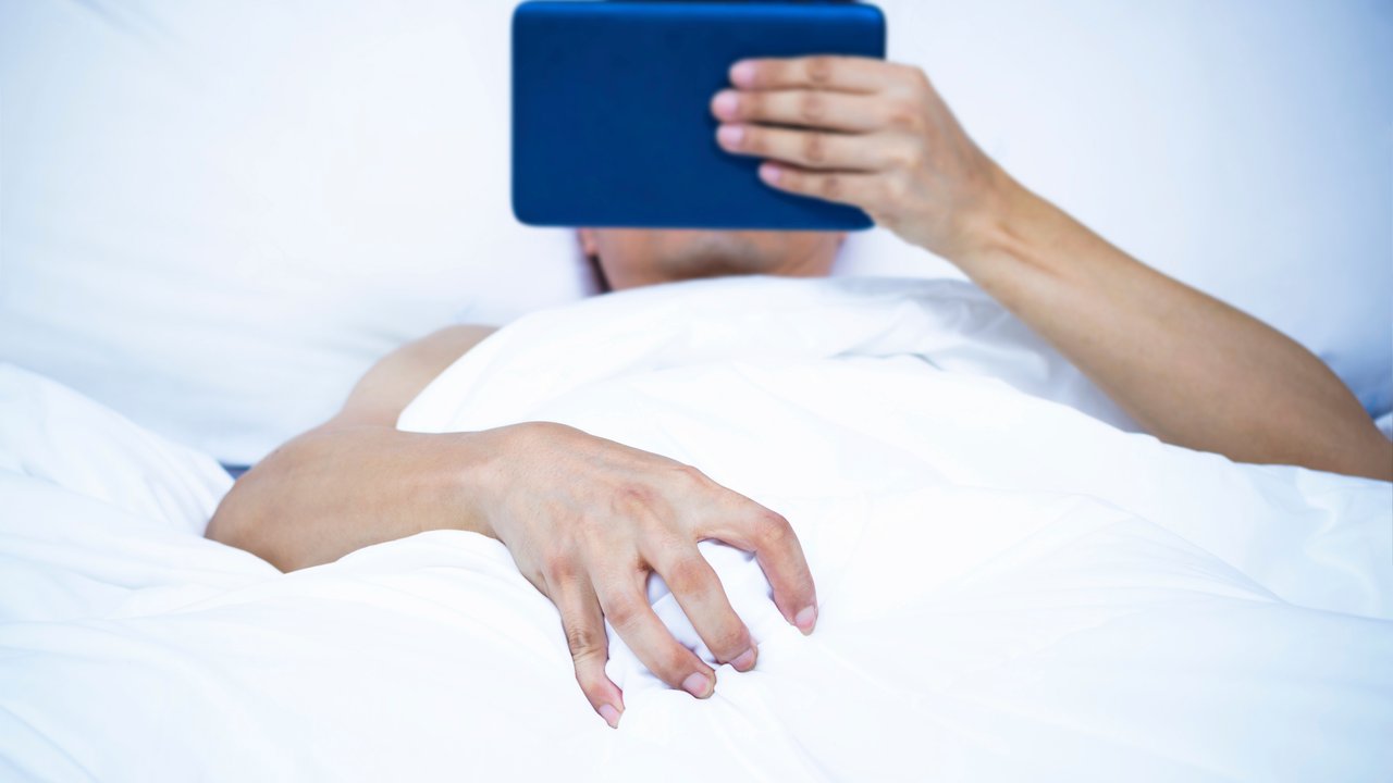 Young asian man lying on white bed &amp; watching pornography on computer tablet. focus to right hand