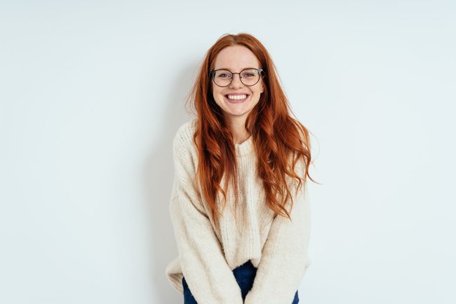Smiling friendly young woman with long red hair wearing spectacles looking at the camera with a vivacious smile against a white interior wall with copy space Smiling friendly young woman with long red hair wearing spectacles looking at the camera with a vivacious smile against a white interior wall with copy space