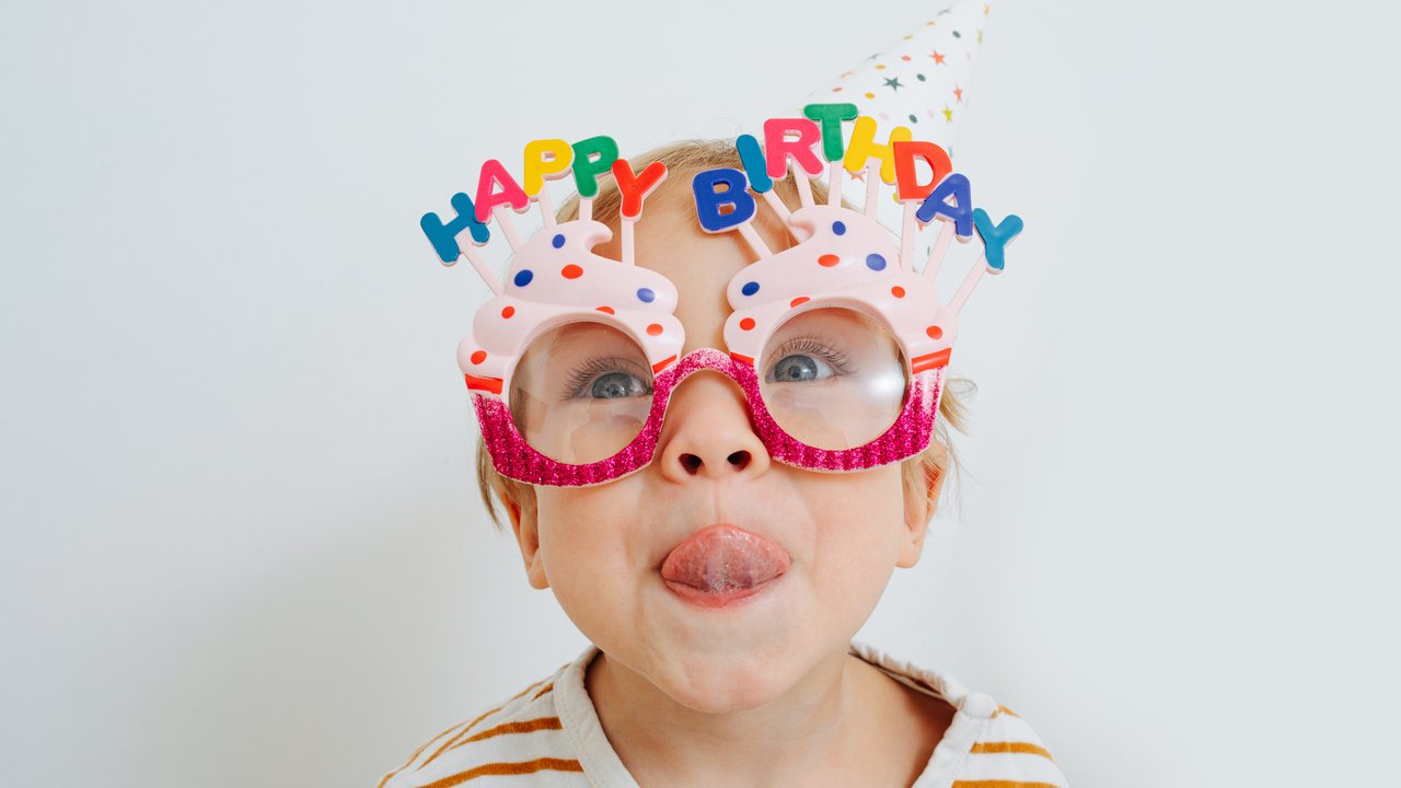 Cute little toddler boy in happy birthday glasses and party hat sticking his tongue out over white.