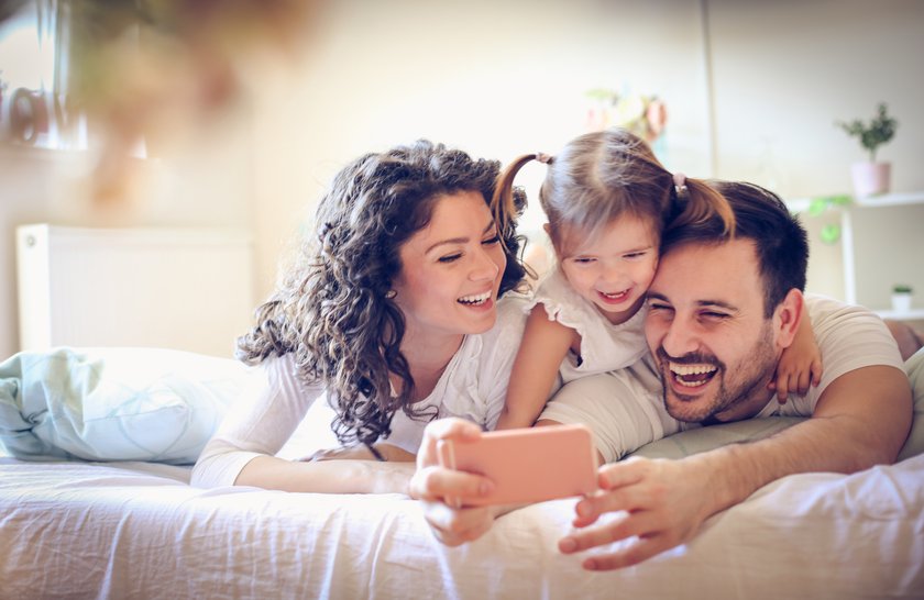 Paar liegt zusammen mit seiner kleinen Tochter im Bett, alle lachen, Vater macht mit seinem Handy ein Foto