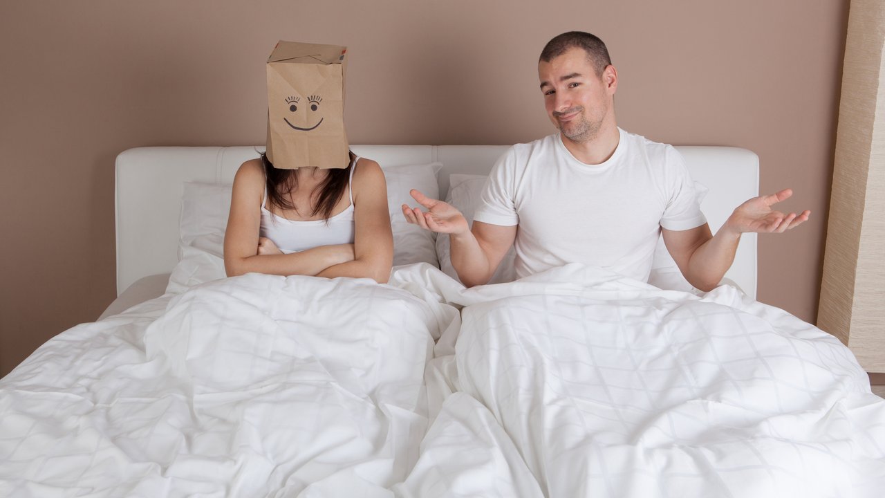 Funny situation in bed. Young couple lying in bed and woman with paper bag over head