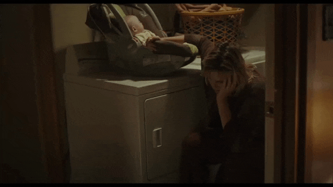Frau sitzt erschöpft und müde vor der Waschmaschine und wippt ihr Baby im Autositz