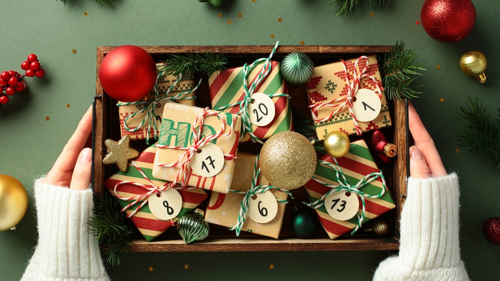 Christmas gifts with numbered advent calendar tags in a wooden box in women's hands on a green background with festive Xmas ornaments and fir branches Christmas gifts with numbered advent calendar tags in a wooden box in women's hands on a green background with festive Xmas ornaments and fir branches