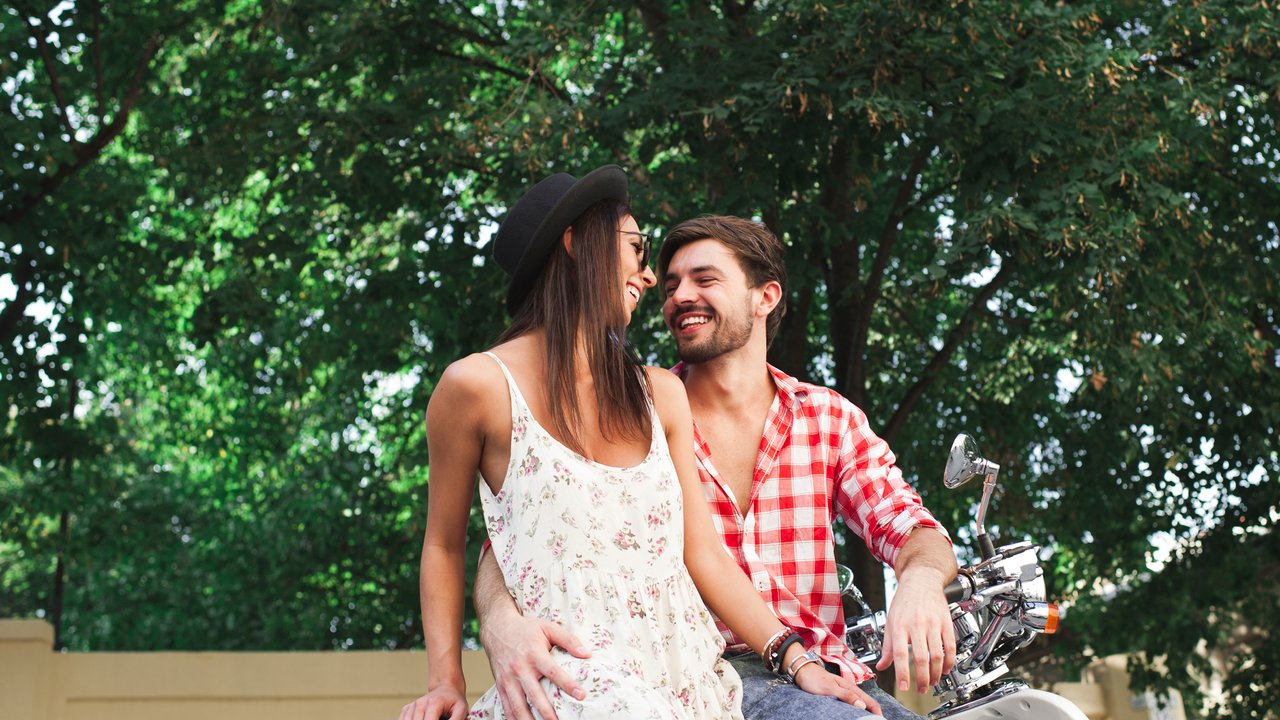 Close up hipster young couple smiling embracing while sitting on a scooter on a summer day on trees background outdoors