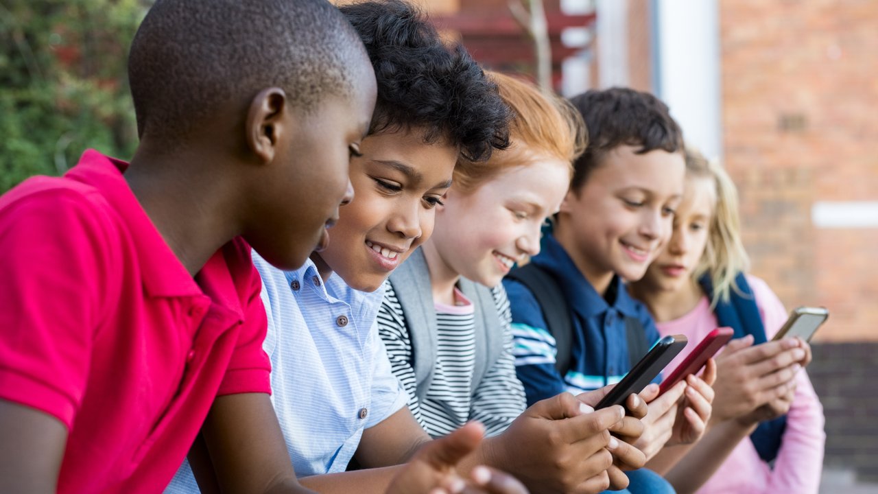 Pupils using mobile phone at the elementary school during recreation time. Group of multiethnic children sitting in a row and typing a message on smartphone. Young boys and girls playing with cellphone.
