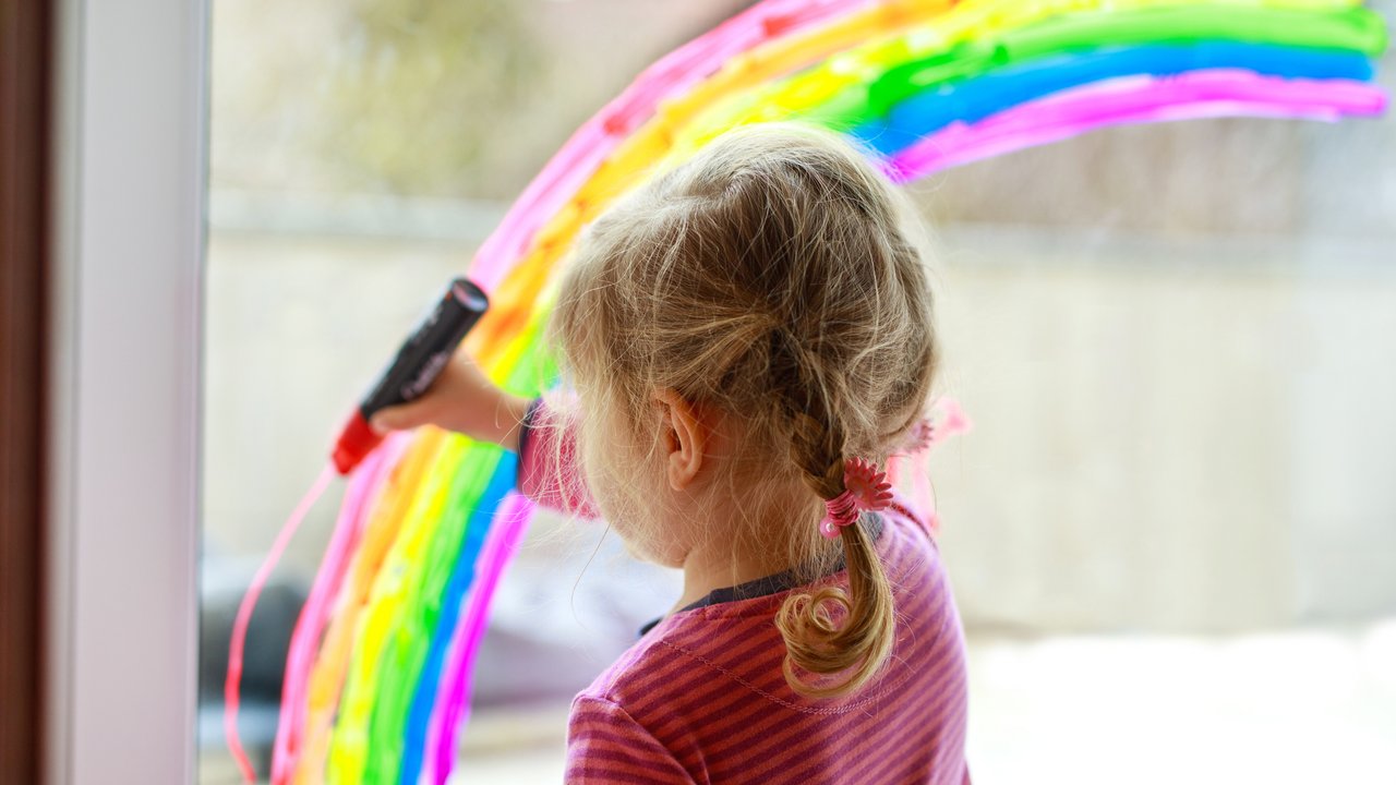 Adoralbe little toddler girl with rainbow painted with colorful window color during pandemic coronavirus quarantine. Child painting rainbows around the world with the words Let's all be well