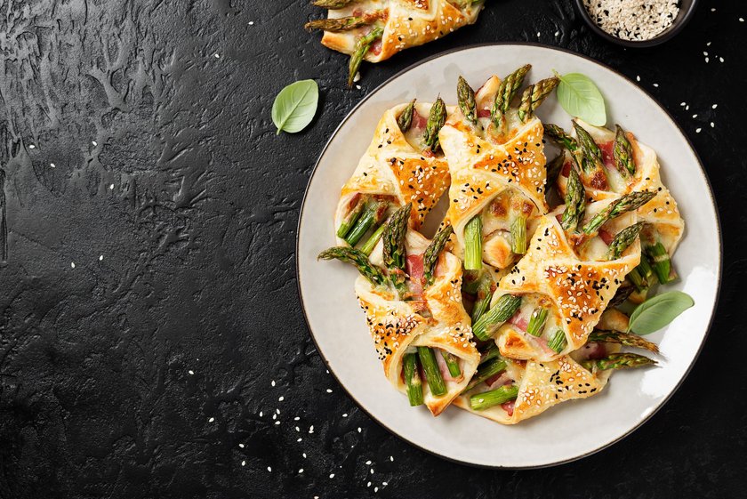 Baked green asparagus with ham and cheese in puff pastry sprinkled with sesame seeds and green basil leaves. Black stone background, top view