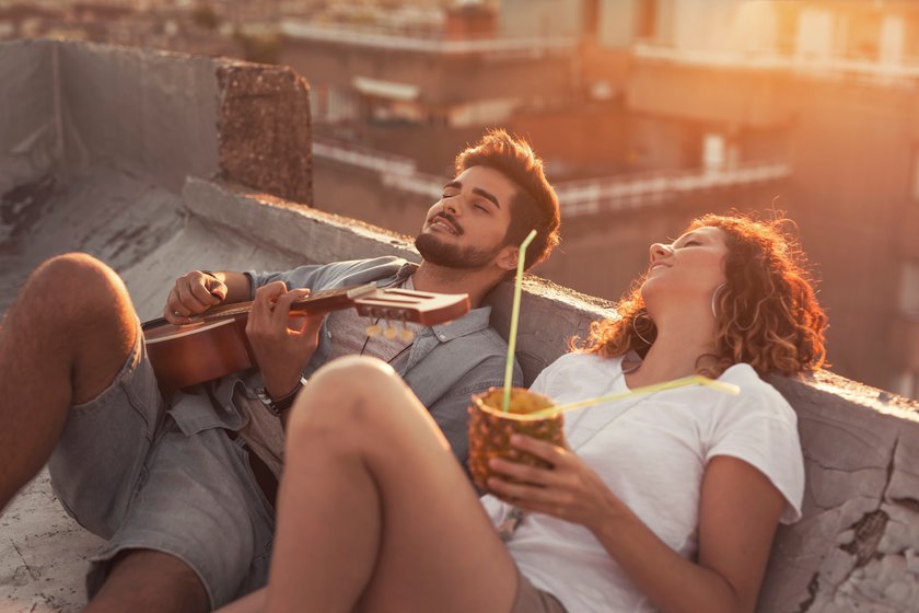 Paar sitzt bei Sonnenuntergang auf einer Dachterrasse, sie hält einen Cocktail in der Hand, er spielt Gitarre