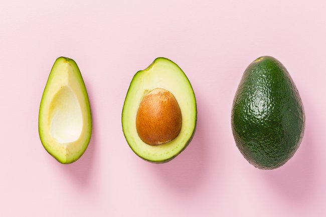 Reife Avocado – ganze und geschnittene frische grüne Frucht mit Samen auf pastellfarbenem, strukturiertem Hintergrund. 