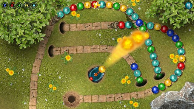 Ansicht eines Levels im Spiel "Marbles Garden". Es zeigt einen grüner Rasen mit Steinpfaden, auf dem bunte Kugeln rollen. Der Spieler steuert eine Kanone, um die farbigen Kugeln zu treffen und so Gruppen von mindestens drei gleichen Kugeln zu bilden, die vom Spielfeld entfernt werden. Die Murmelkanone schießt in diesem Bild mit einem Spezialbooster Feuer ab. Im Hintergrund sind Bäume und Blumen zu sehen. 