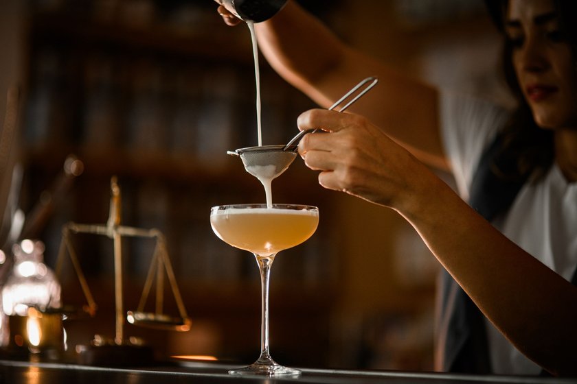 Female bartender straining frothy champagne-colored cocktail through a sieve into a stemmed glass, holding shaker high above bar counter on blurred background