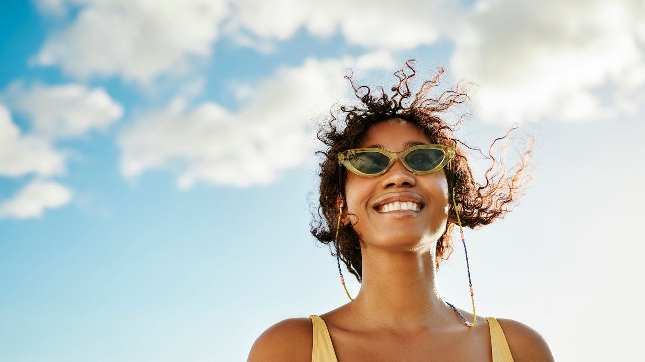 Eine junge Frau, die einen modischen Badeanzug und Sonnenbrillen trägt, lächelt strahlend, während sie draußen unter einem klaren Sommerhimmel steht.