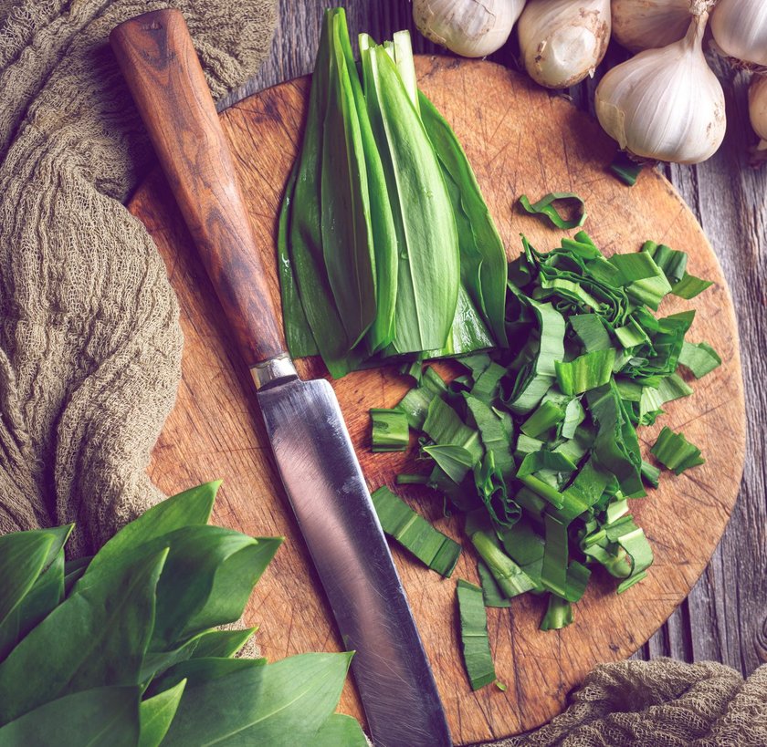 Cutting fresh organic bear's wild garlic leaves on wooden board with knife close up. Food photography