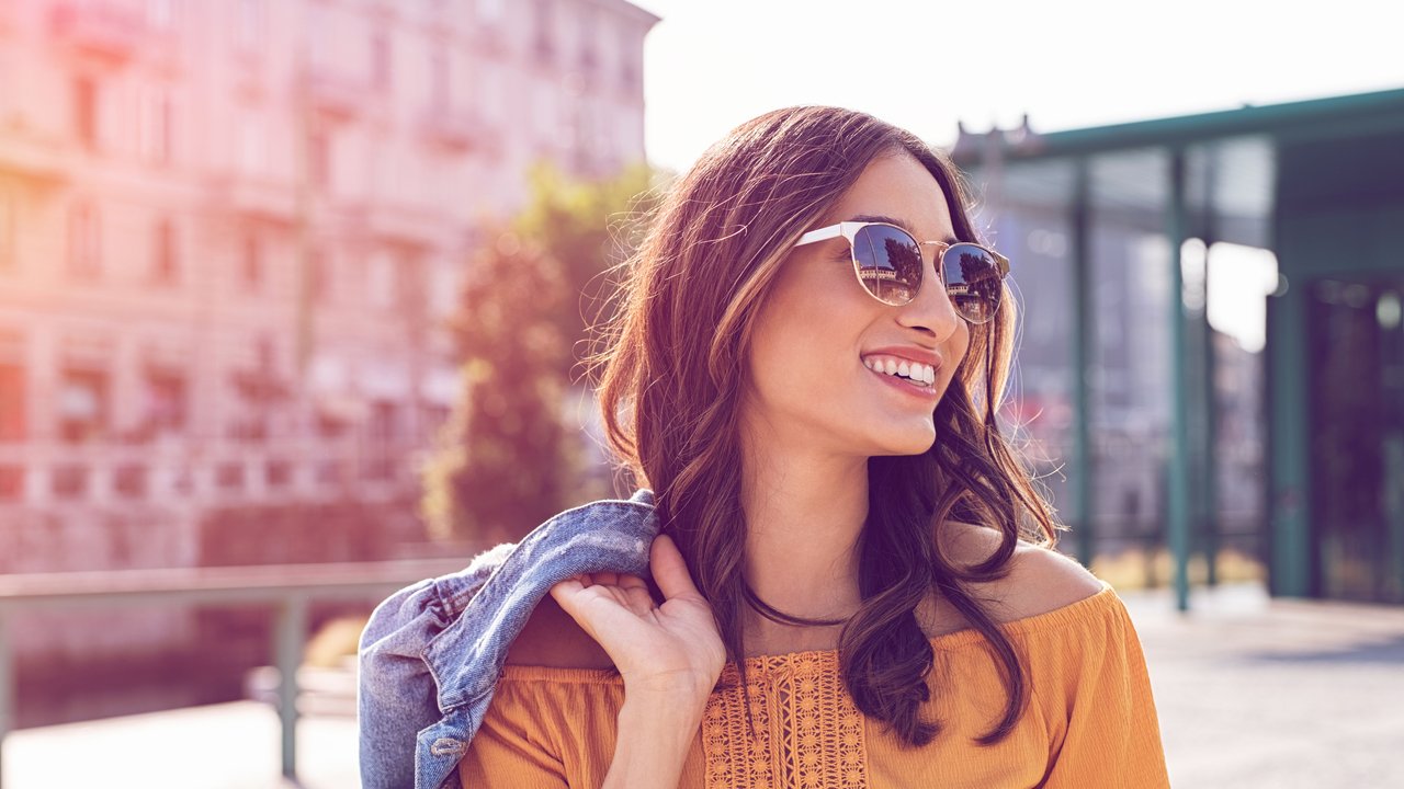 Happy latin woman holding denim jacket and walking on the street on a bright sunny day. Cheerful stylish girl with sunglasses enjoying the spring. Young woman wearing shades while looking away in the afternoon.