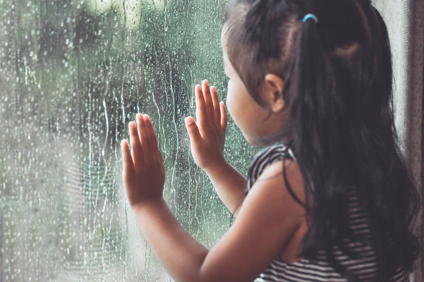 Kind steht am Fenster und schaut sich draußen den Regen an
