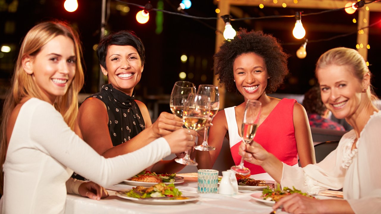 Group of female friends eating dinner at rooftop restaurant, holding glasses of wine, smiling to camera