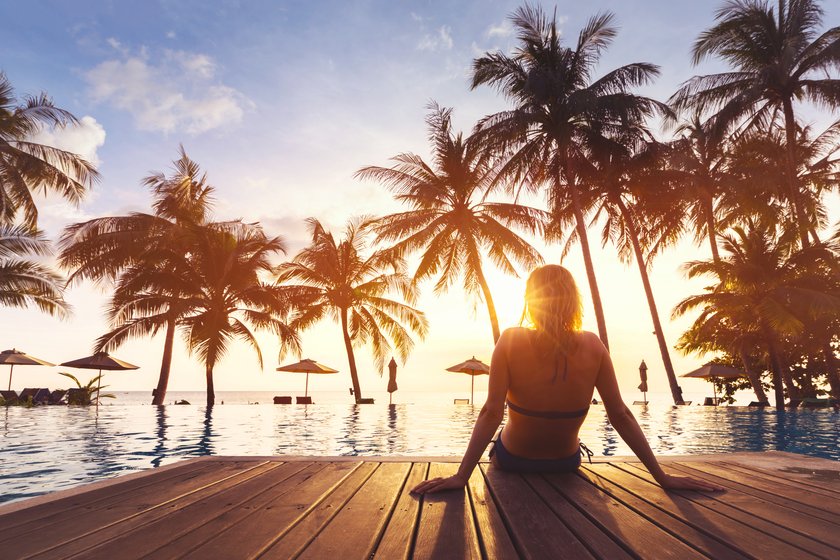 Frau sitzt bei Sonnenuntergang am Pool und schaut auf Palmen