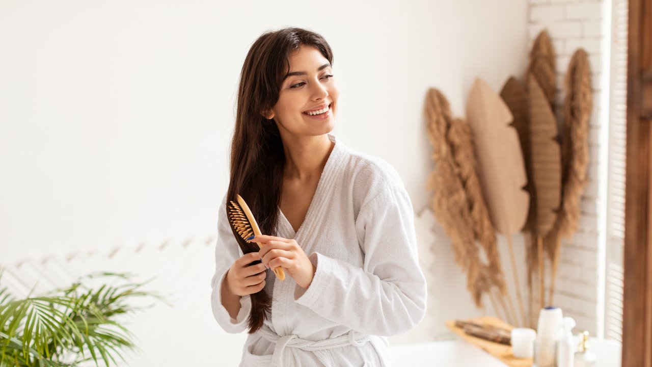 Haircare Concept. Brunette Woman Brushing Smooth Hair With Wooden Hairbrush Smiling Looking Aside Posing Wearing Dressing Gown In Modern Bathroom At Home. Hair Detangling And Beauty Routine