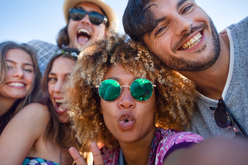 Group of friends having fun taking a selfie. They are all looking at the camera and smiling and laughing. Some are making funny faces. Background is blue sky on a sunny day.