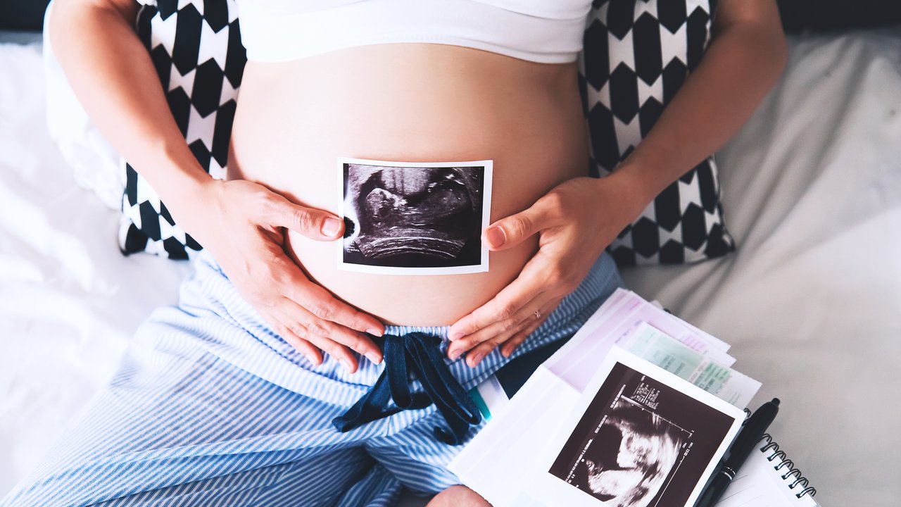 Pregnant woman makes notes in notebook and holding ultrasound image and medical documents at home interiors. Pregnancy, parenthood, preparation and expectation concept. Close-up, copy space, indoors.