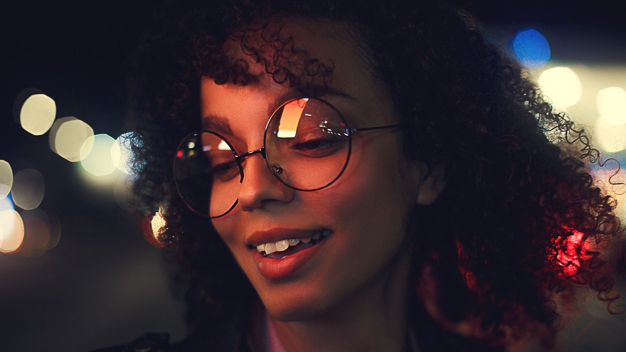Portr&#x00E4;t&#x20;einer&#x20;jungen&#x20;Frau&#x20;mit&#x20;lockigem&#x20;Haar&#x20;und&#x20;Brille.&#x20;Es&#x20;ist&#x20;nachts&#x20;und&#x20;es&#x20;leuchten&#x20;Lichter