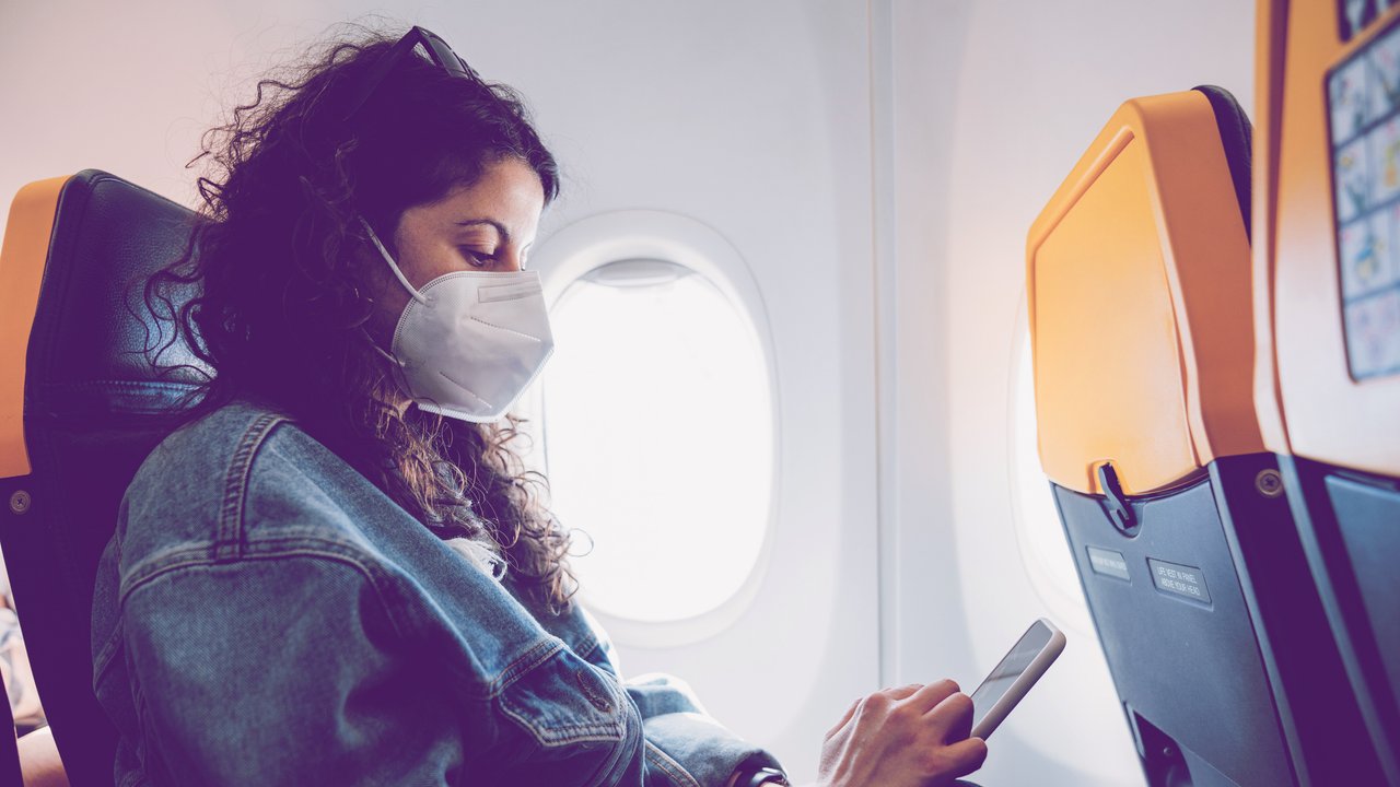 Woman on an airplane is texting on her smart phone. She's wearing a protective face mask.