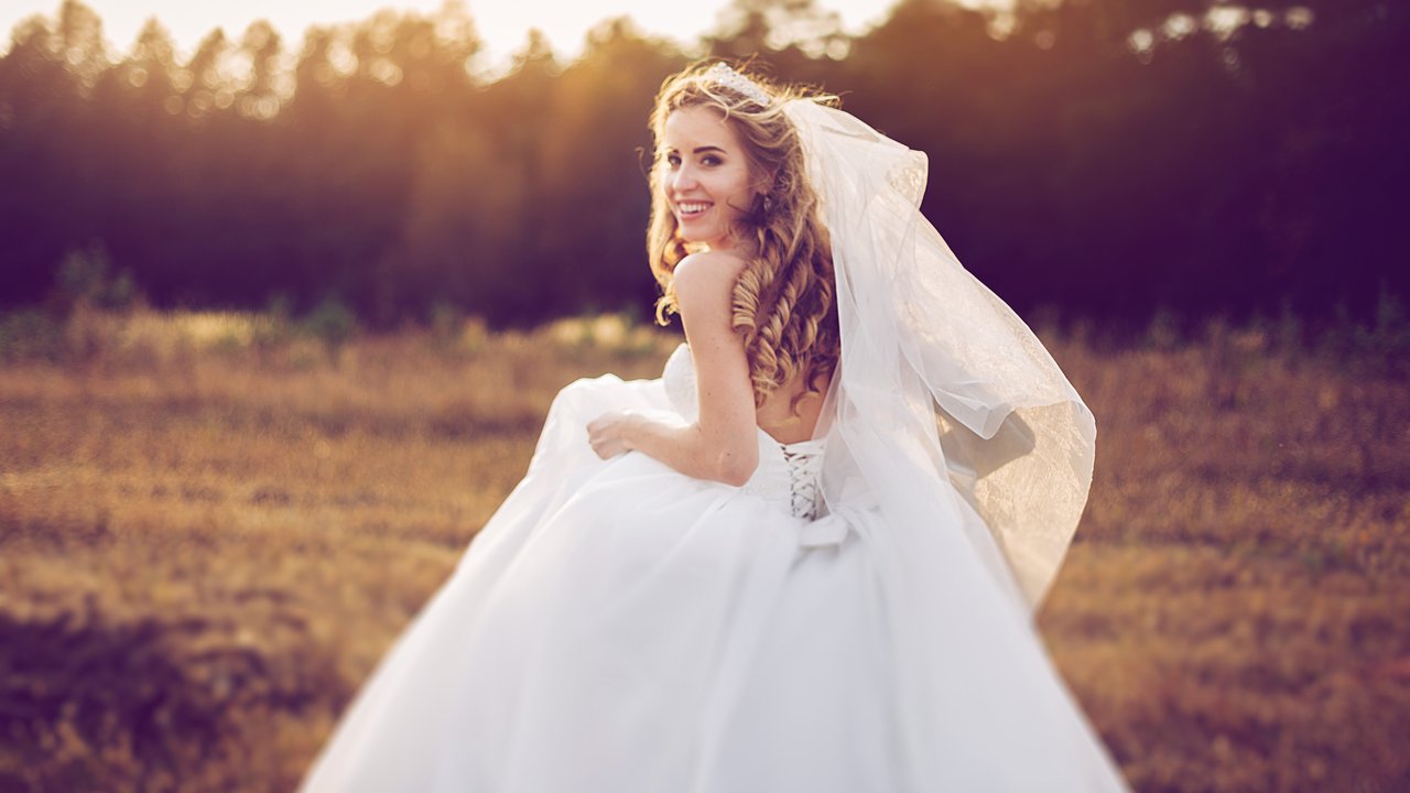 Nice portrait of a beautiful bride with a diadem on her head. She is running in the field, holding her wedding dress in her hands and looking at the camera.