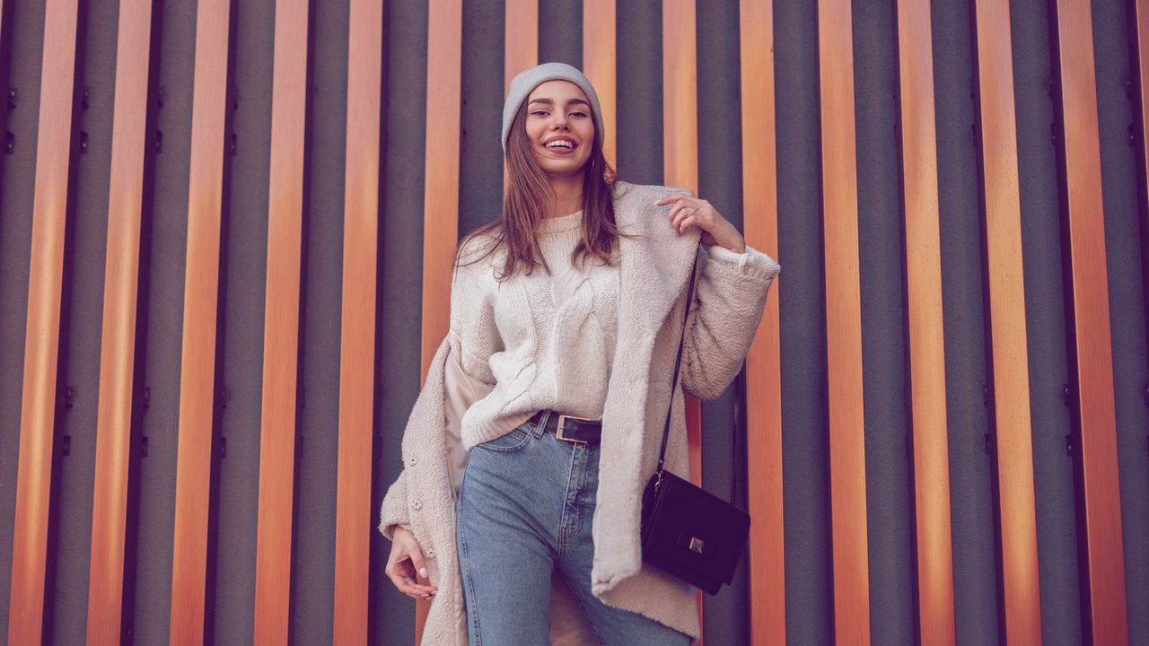 Pretty young happy woman in a warm sweater in a knitted hat in a fashionable faux fur coat in vintage jeans with a leather black handbag stands and smiles near a metal wall. Positive girl model.