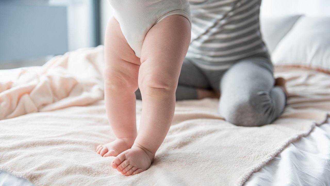 Close up of infant legs standing on bed, female sitting on background. Focus on feet