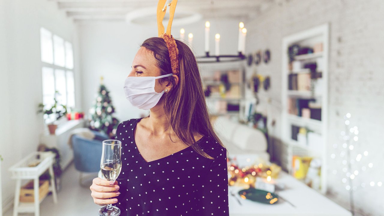 Photo of a young woman holding a glass of champagne, wearing a protective face mask and reindeer antlers; celebrating and hosting a Christmas dinner party at home during the COVID-19 pandemic.