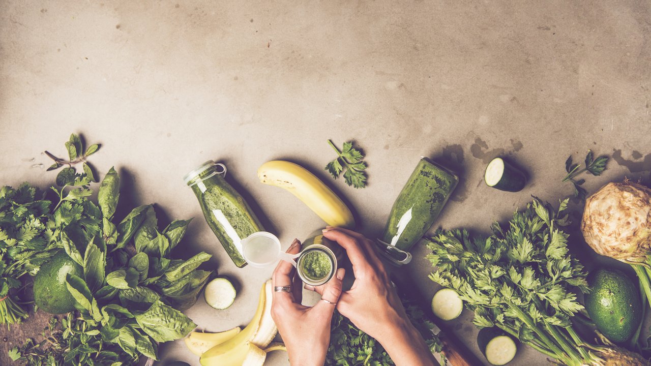 Making green detox smoothie. Flat-lay of ingredients for making smoothie drink and female hands with bottle over concrete background, top view, copy space, wide composition. Clean eating food concept