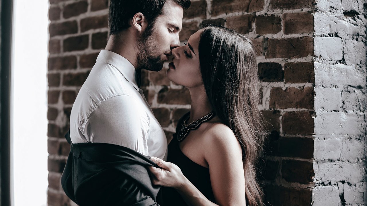 Close-up side view of beautiful young woman in cocktail dress kissing her boyfriend and taking off his jacket while standing near brick wall in loft interior