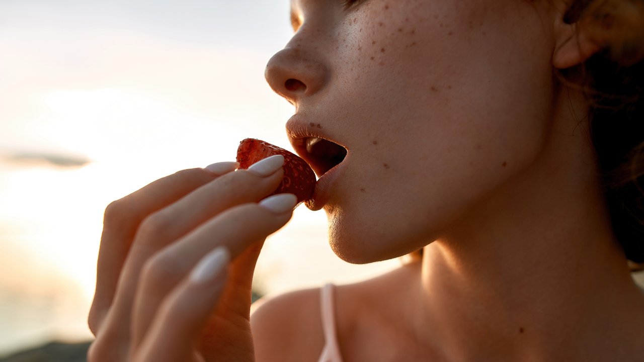 Close up view of young caucasian girl lips licking strawberry while resting on beach in evening, widescreen, cropped. Romantic date concept