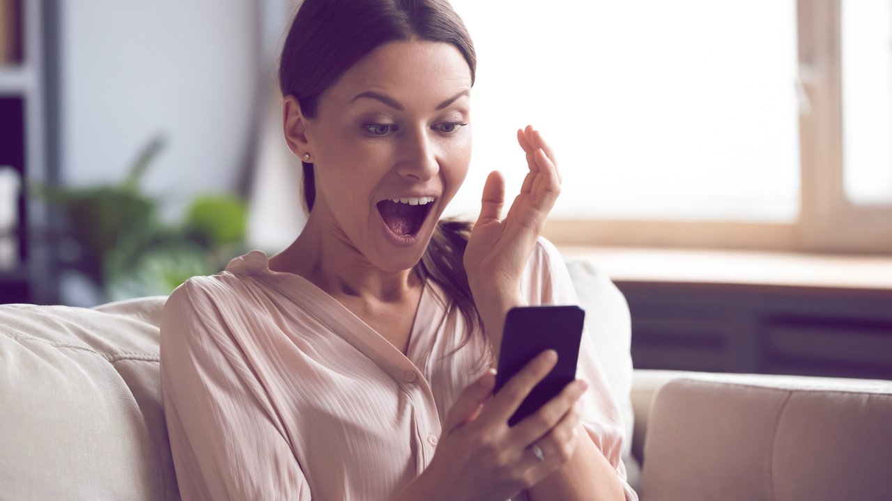 Amazed young woman sit on couch at home feel shocked reading unexpected good news on cellphone gadget, stunned millennial girl surprised by online lottery win on smartphone, luck concept