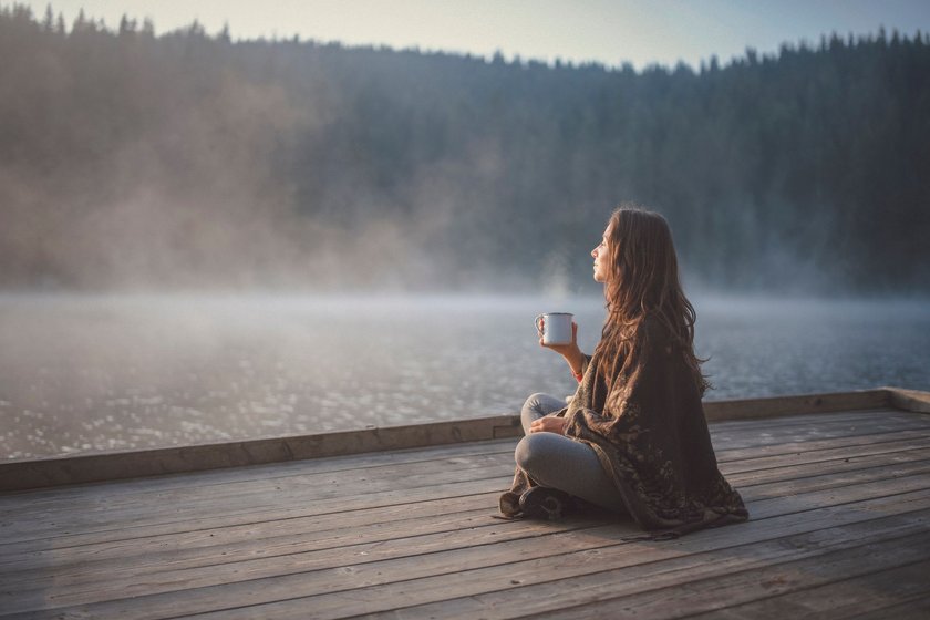 Seitenansicht einer Frau, die auf einem hölzernen Steg in der Natur sitzt, den Sonnenaufgang betrachtet und Kaffee trinkt.