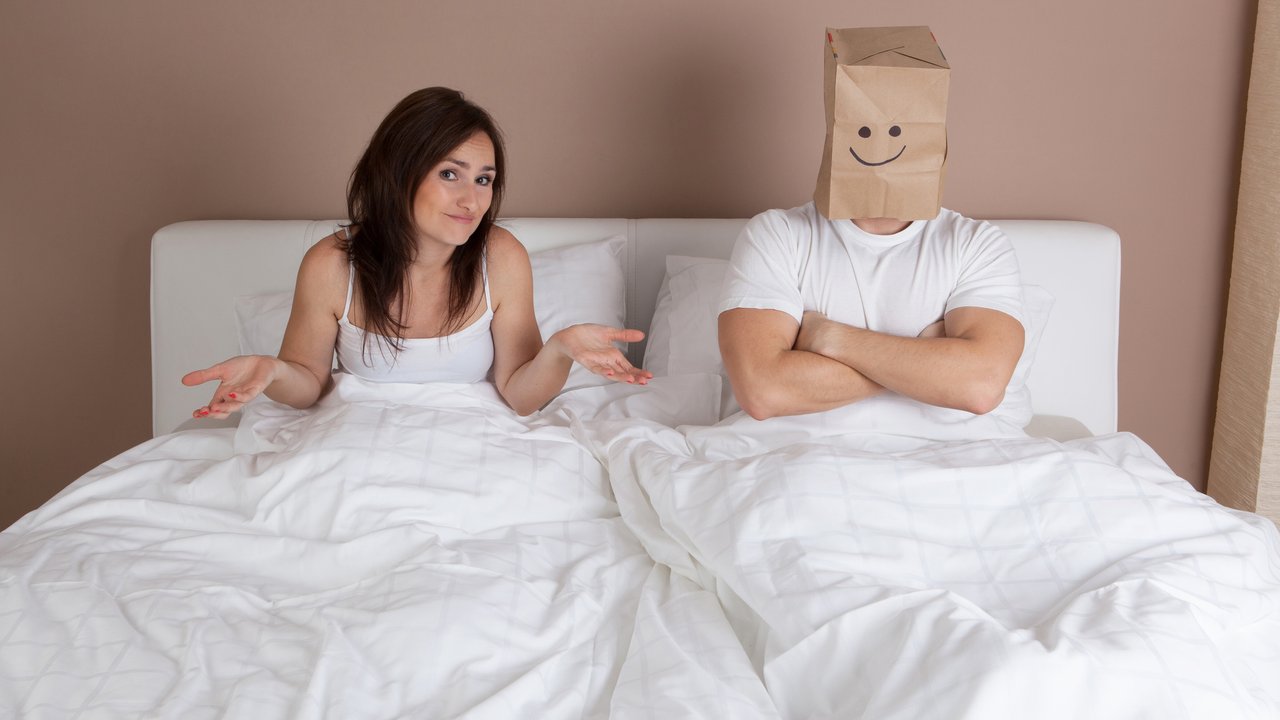 Funny situation in bed. Young couple lying in bed and man with paper bag over head