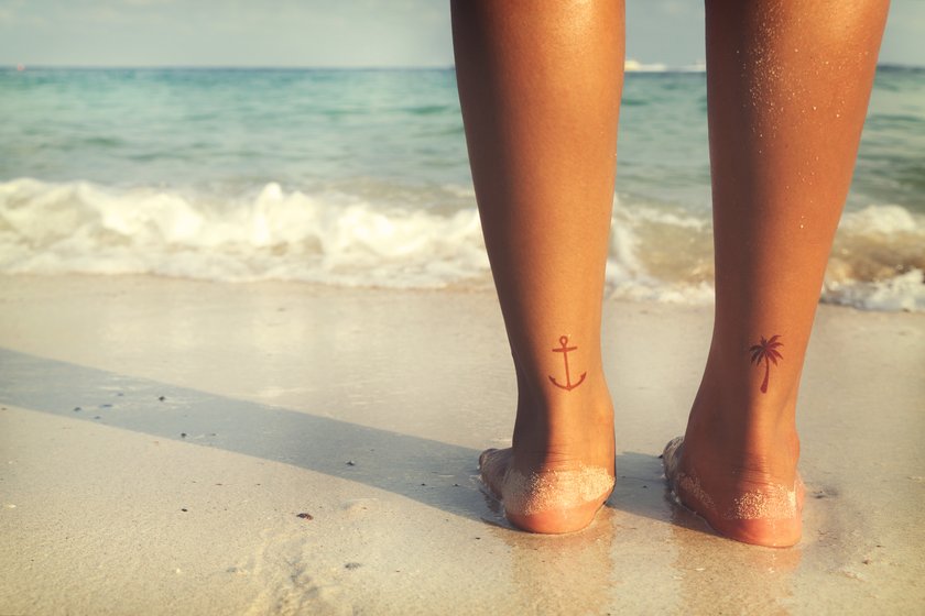 Frau am steht am Strand, auf ihren Fersen hat sie ein Anker- und ein Palmen-Tattoo