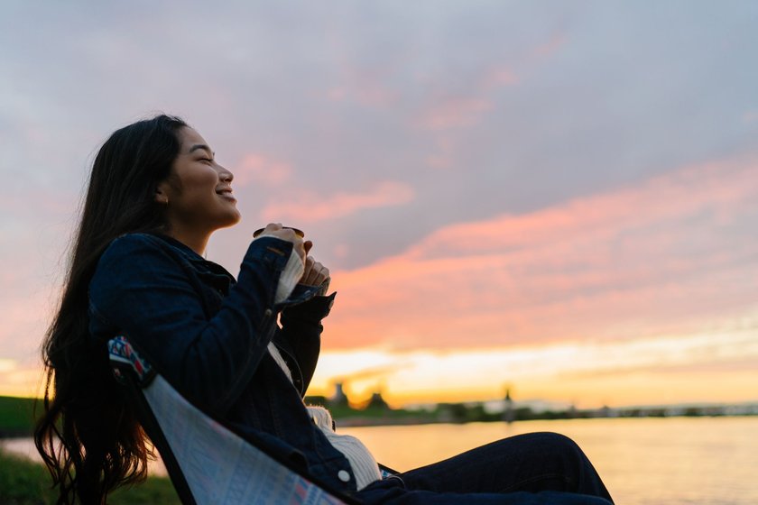 Eine junge Frau genießt es, in der Natur am See zu sitzen und während des Sonnenuntergangs ein heißes Getränk zu trinken